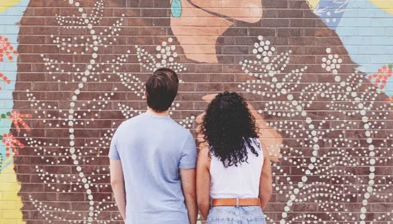 couple admiring painted mural "ᑮᐦᑳᔮᓱᐁᐧᐤ KÎHKÂYÂSOWÊW / She Shines Brightly"