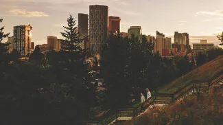 two friends walk down an outdoor staircase lined with trees. the downtown calgary skyline is prominent in the background.