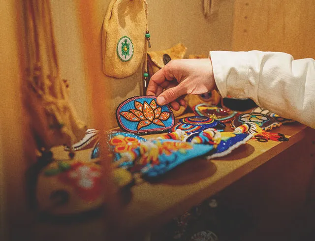 Someone picks up a Beaded Orange Flower Barrette from a shelf full of other beadwork arts and crafts as well as ceremonial and medicine bags