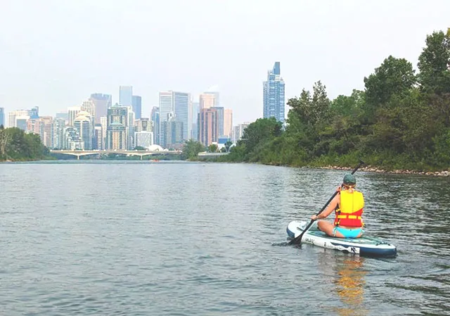 woman paddle boarding down the Bow River with the city skyline in the background