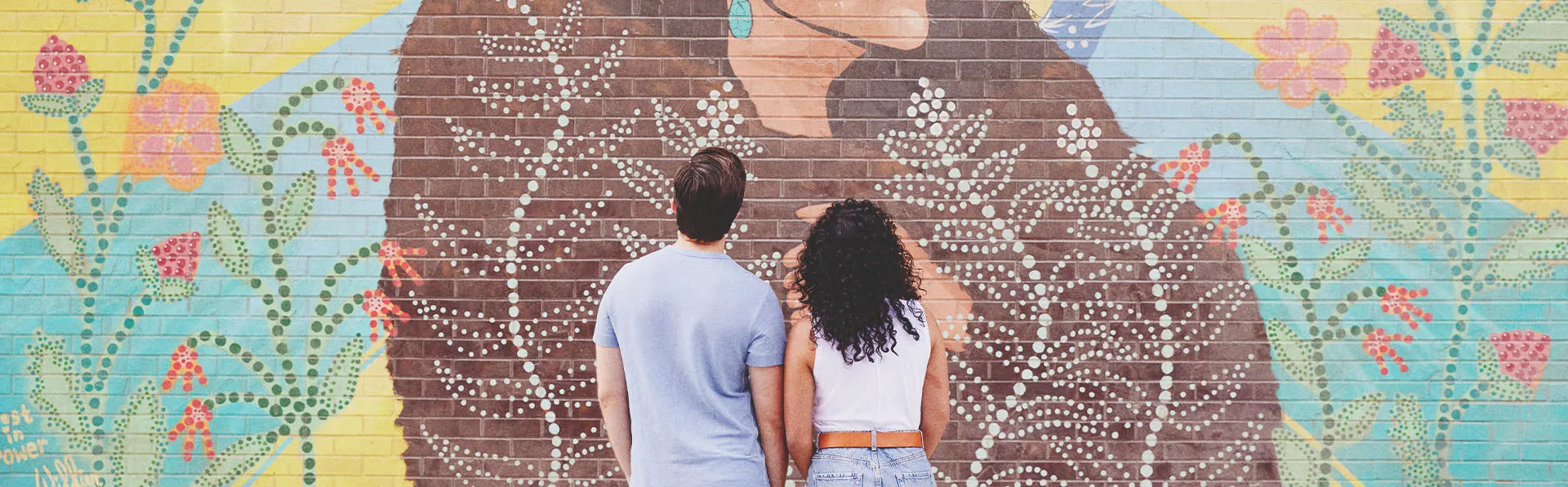 couple admiring painted mural "ᑮᐦᑳᔮᓱᐁᐧᐤ KÎHKÂYÂSOWÊW / She Shines Brightly"