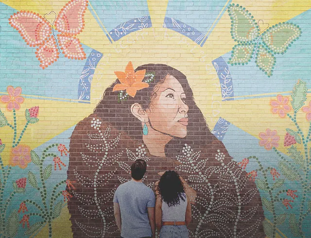 couple admiring painted mural "ᑮᐦᑳᔮᓱᐁᐧᐤ KÎHKÂYÂSOWÊW / She Shines Brightly"